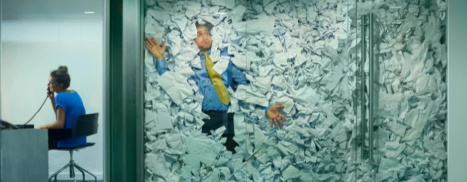A businessman is trapped behind a glass office door, buried under an avalanche of discarded paper. In the adjacent office, a colleague works on, completely unaware. A visual metaphor for information overload, fragmented systems, and the chaos of unmanaged knowledge in the modern workplace.