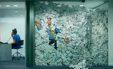 A businessman is trapped behind a glass office door, buried under an avalanche of discarded paper. In the adjacent office, a colleague works on, completely unaware. A visual metaphor for information overload, fragmented systems, and the chaos of unmanaged knowledge in the modern workplace.