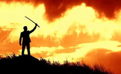Silhouette of person in a suit holding. sword against an amber sunset. 